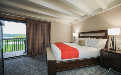 Picture of a lodge room. There is a bed with white sheets and a red blanket and through the window you can see a deck.